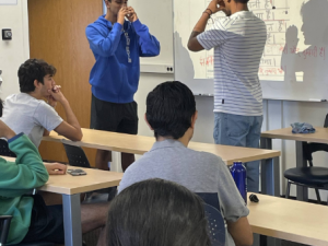 Two Duke Hindi students making shapes with their hands to perform their poems through action in Dr. Harsh Bola's poetry workshop in Hindi class. Other students are sitting at desks watching the performance.