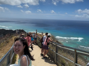 Jiani (Thea) Yu posing on a boardwalk in Hawaii with other visitors and the ocean pictured in the background.