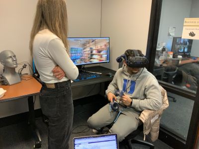 A student wearing a grey Duke sweatshirt sitting down wearing a virtual reality (VR) headset. The student is also holding a controller in their hands. In the background there is another person watching the student as well as a computer monitor displaying what the student using VR is seeing (the inside of a store with clothes displayed on shelves).